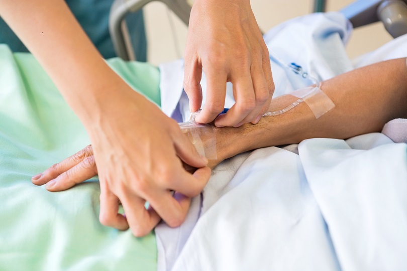 Foto ilustrativa das mãos de uma enfermeira e de uma paciente. A enfermeira está colocando o acesso intravenoso no dorso da mão esquerda da paciente.