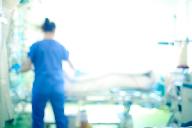 Foto ilustrativa mostra imagem desfocada de profissional da saúde em frente a um leito hospitalar. Ela está de costas e usa uniforme azul claro.