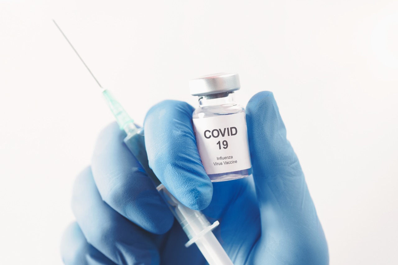Foto de uma mão com luva segurando uma ampola de vacina e uma seringa. No vidro da ampola está escrito "Covid-19"