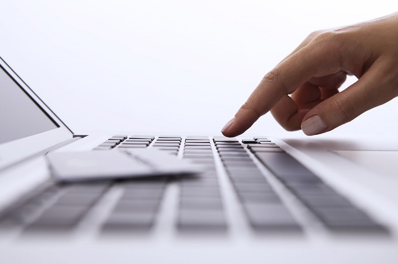 Foto ilustrativa de um notebook sendo visualizado de perfil. Na foto também se vê  um dedo da mão de uma mulher apertando uma tecla do teclado.