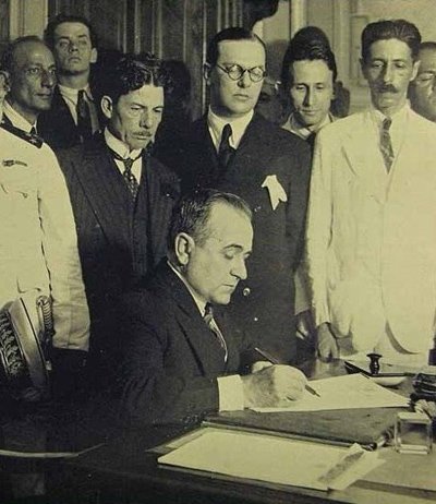 Foto de Getúlio Vargas, sentado assinando a nomeação de ministros. Atrás dele, em pé, aparecem seis homens em traje formal. Um dos que está mais próximo de Getúlio é Lindolfo Collor, primeiro ministro do Trabalho, Indústria e Comércio