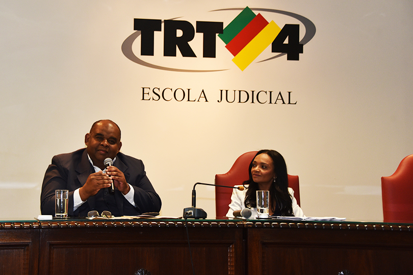 Fotografia da juíza Karen Luise Vilanova Batista de Souza Pinheiro e do advogado Osvaldo Ferreira Reis, sentados à mesa durante palestra, com logotipo do TRT-RS ao fundo