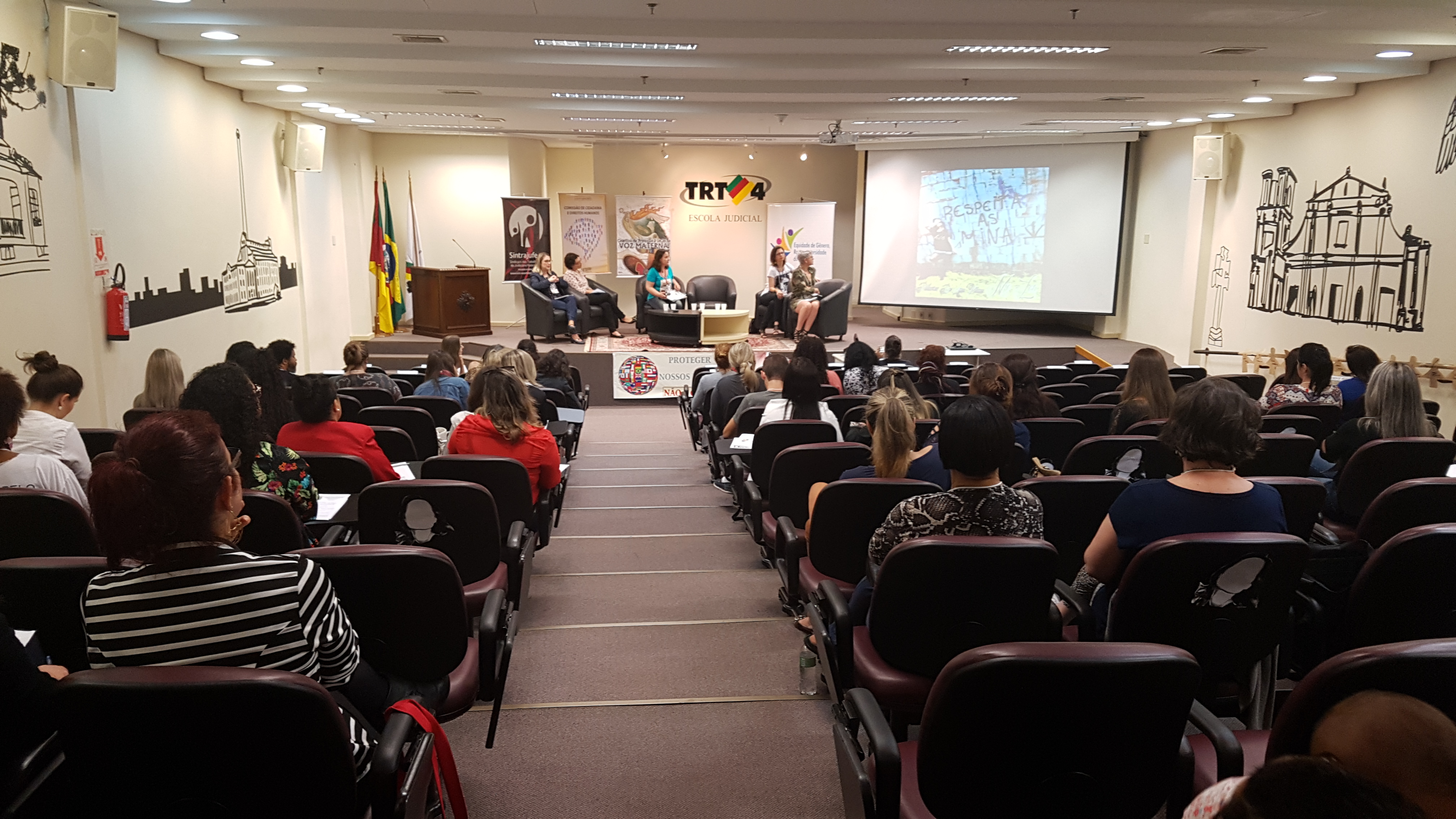 Fotografia mostra plateia e painelistas sentadas em poltronas no palco do auditório ao fundo