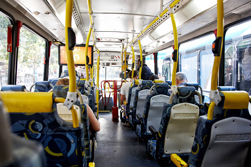 Imagem de interior de ônibus, com bancos, alguns passageiros e o cobrador aparecendo ao fundo.