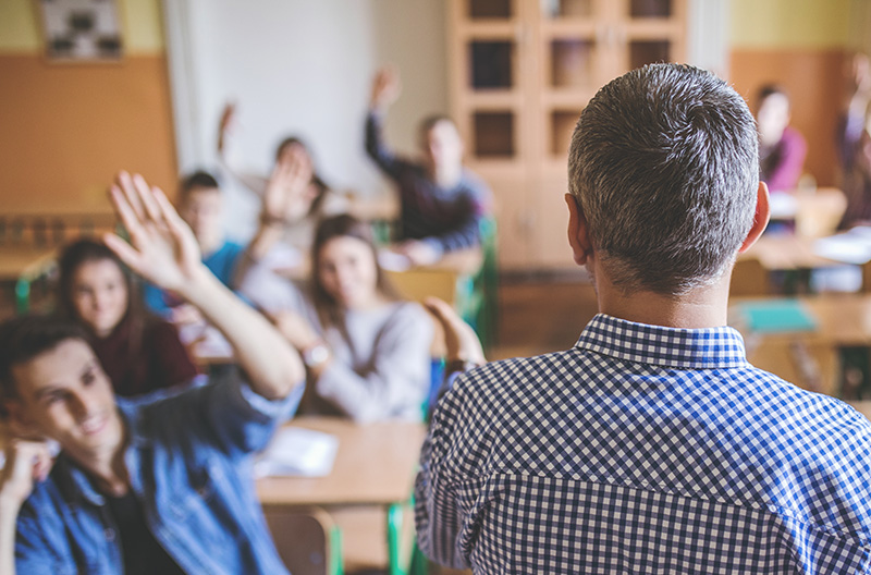 Professor de costas, virado para os alunos adultos, alguns dos quais com as mãos levantadas.