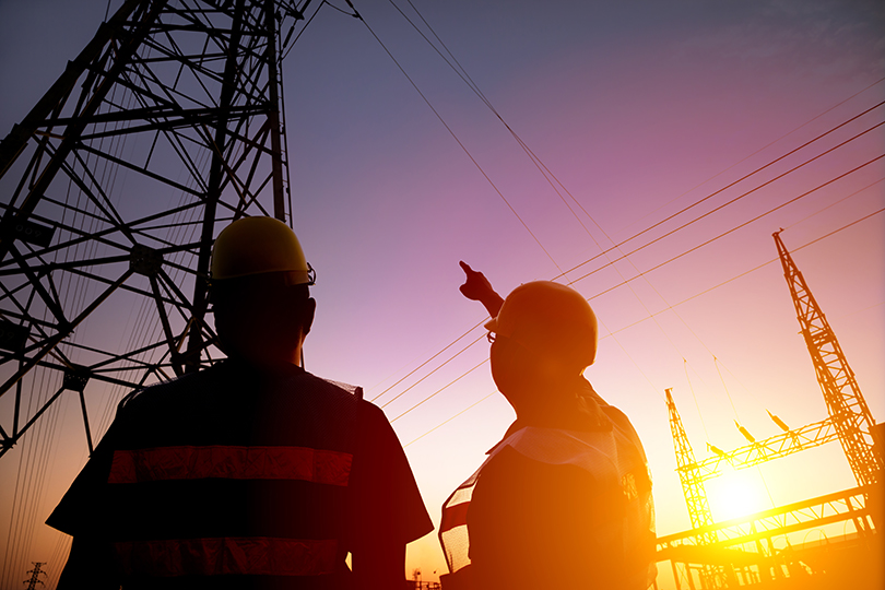 Fotografia de dois homens vestindo capacetes de proteção olhando para uma torre de transmissão de energia elétrica, um deles apontando o dedo na direção da torre.