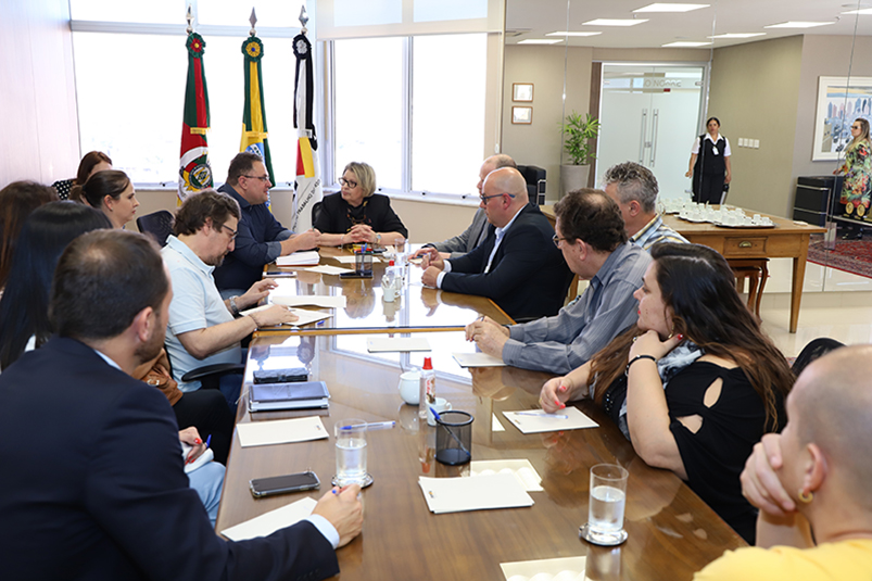 Mesa de reuniões cercada de pessoas no Fórum de Relações Institucionais. Presidente do TRT-RS, desembargadora Vania Cunha Mattos, na ponta da mesa, ao fundo da fotografia.