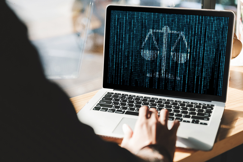 fotografia em que um homem mexe em um computador em cuja tela aparece a imagem de uma balança formada por zeros e uns fazendo alisão à linguagem binária dos equipamentos eletrônicos