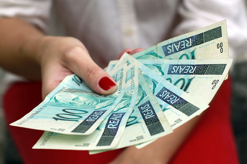 Mãos femininas segurando notas de 100 reais.