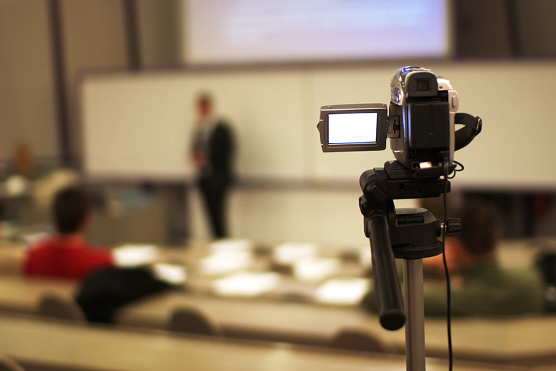 Fotografia de uma câmera, em primeiro plano, gravando um professor dando aula ao fundo, desfocado.