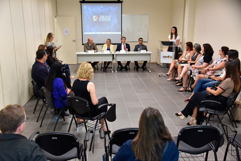 Imagem aberta da cerimônia, mostrando a mesa oficial e os servidores formandos, sentados.