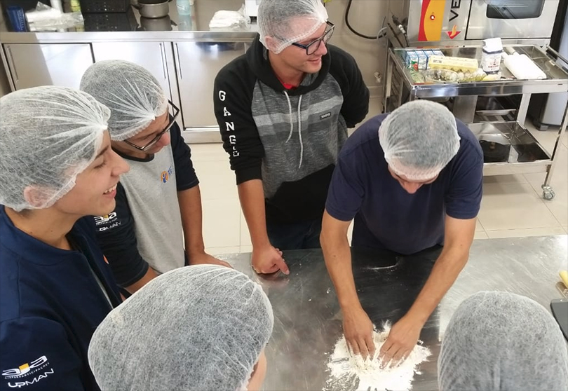 Um grupo de seis alunos na cozinha. Um deles está trabalhando com a massa e outros observam sorridentes.