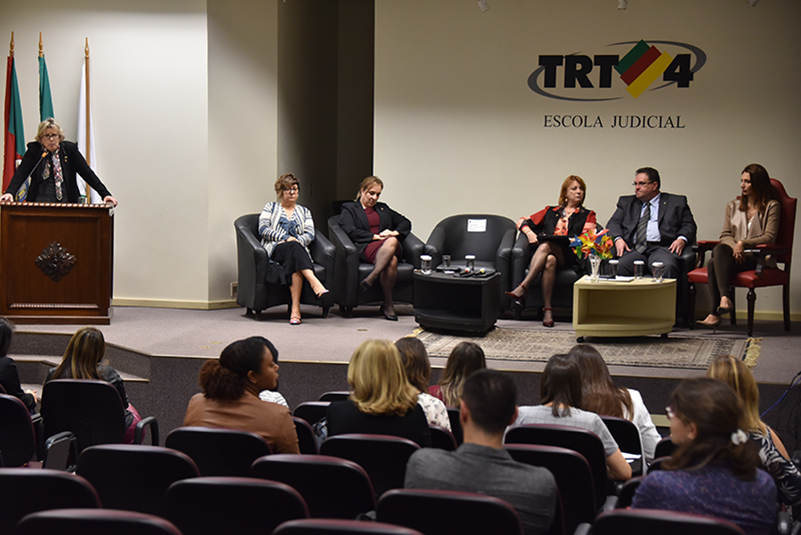 Imagem aberta do palco do auditório, aparecendo os debatedores sentados e, à esquerda, a presidente Vania Mattos fazendo seu pronunciamento, no púlpito.