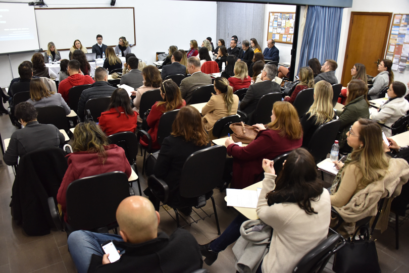 Fotografia do evento com plateia em primeiro plano e, ao fundo, os palestrantes sentados à mesa.