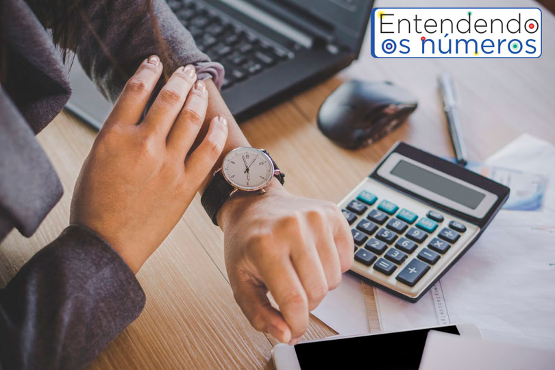 Imagem de um homem sentado à uma mesa de trabalho com notebook, calculadora e documentos. Ele está olhando as horas em um relógio de pulso. A foto leva, também, a logomarca da editoria "Entendendo os Números".