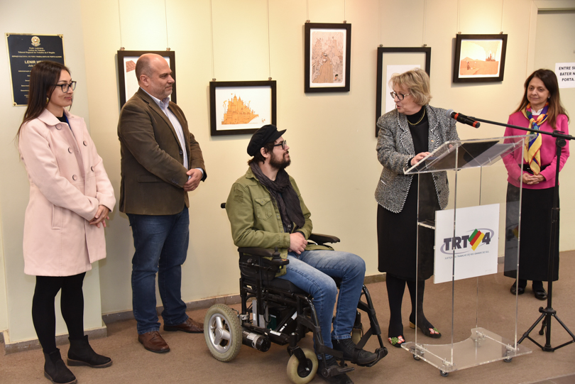 Foto tirada durante cerimônia de abertura. Aparecem a presidente Vania, fazendo seu pronunciamento no púlpito, ao lado do artista Rafael, do desembargador Lucena, da servidora Milene e da juíza Anita. Ao fundo, os cartuns afixados na parede da galeria.