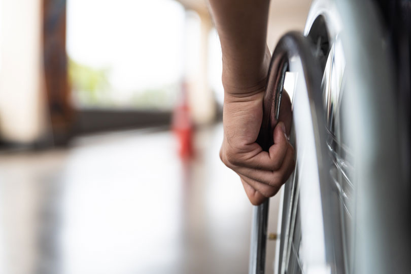 Foto de um cadeirante chegando em um prédio. Ângulo fechado na mão do cadeirante sob a roda da cadeira de rodas. Foto de banco de imagens.