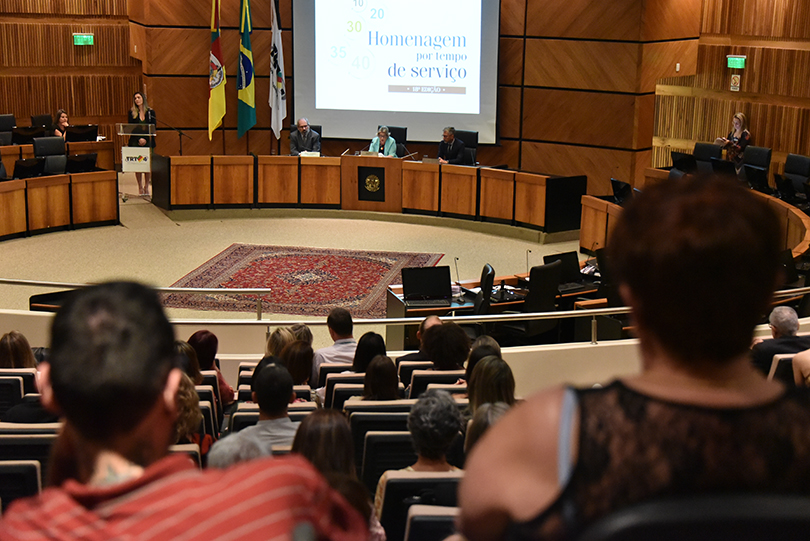 Foto do público em primeiro plano e, ao fundo, a mesa oficial da cerimônia, com a presidente Vania Cunha Mattos, o corregedor Marçal Henri dos Santos Figueiredo e o vice-corregedor Marcelo Gonçalves de Oliveira