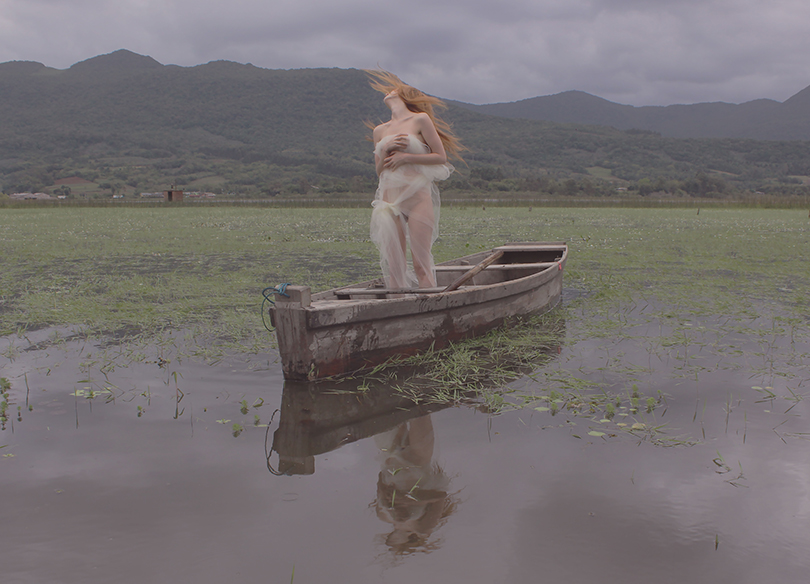 Imagem de uma mulher cuja cabeça fazendo um movimento para trás, com seu corpo nu envolto em um véu translúcido, de pé em uma canoa que está parada na água de uma lagoa, entre juncos, emoldurada pela Serra do Mar e um céu cinzento.