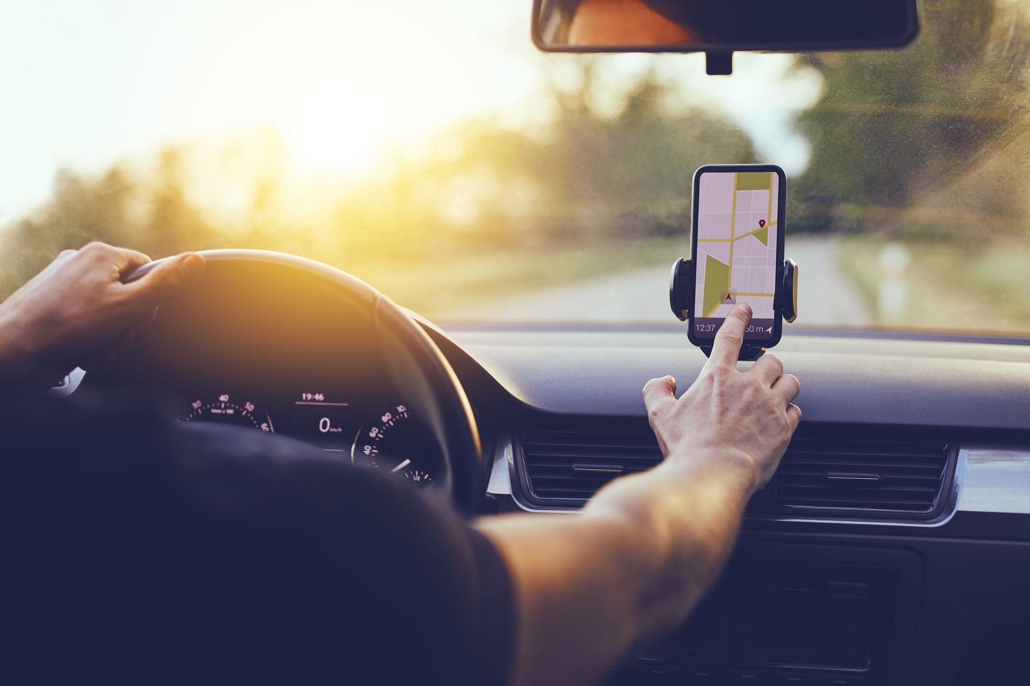 Imagem mostra volante e painel do carro. Acima do painel está um celular no qual o motorista seleciona, em um aplicativo, o mapa do trajeto a ser feito.