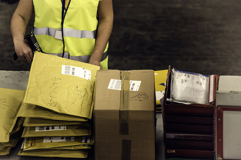 Pra Cego Ver: foto de vm/iStock Banco de Imagens mostra pilha de envelopes e caixas em uma esteira sendo separadas por um trabalhador dos correios.