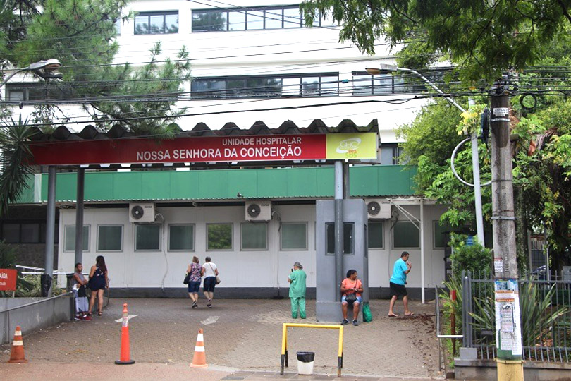 Pra Cego Ver: fachada do Hospital Nossa Senhora da Conceição, com algumas pessoas em frente.