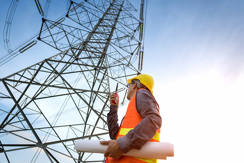 Pra Cego Ver: foto de af_istocker/iStock Banco de Imagens mostra um trabalhador em frente a uma torre de alta tensão.