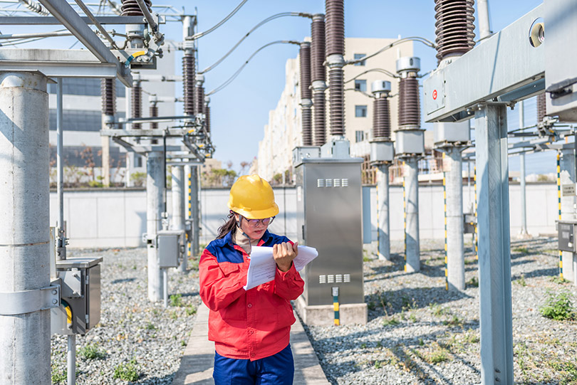 Pra Cego Ver: Foto de Young777/iStock Banco de Imagens mostra mulher fazendo anotações em estação de geração de energia elétrica. Uniforme e capacete dão a ideia de que se trata de uma engenheira eletricista.