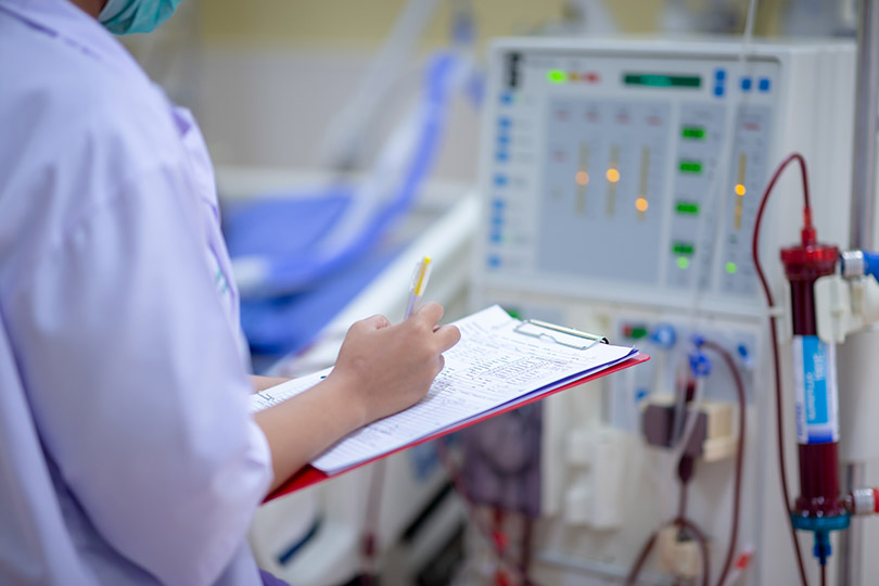 Pra Cego Ver: Foto de Saengsuriya13/iStock Banco de Imagens mostra imagem do braço de um profissional da saúde preenchendo prontuário em frente a uma máquina de hemodiálise.