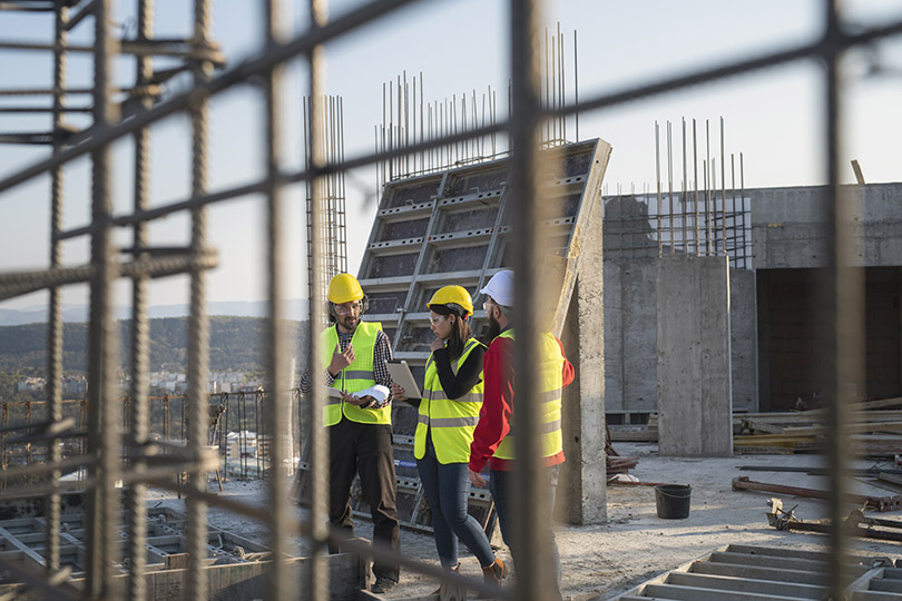 Pra Cego Ver: Foto de ArtistGNDphotography/iStock Banco de Imagens mostra dois engenheiros e uma engenheira em uma obra de construção civil