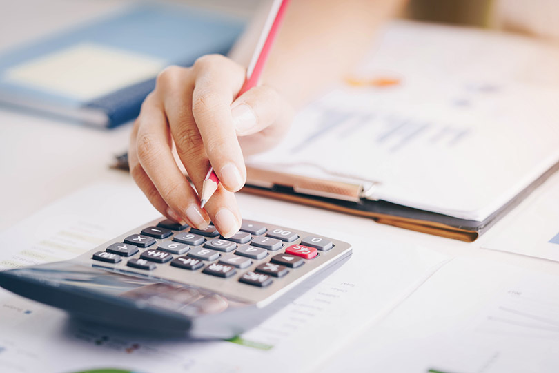 Pra Cego Ver: Foto de Thailand Photographer./iStock Banco de Imagens mostra mão de mulher utilizando calculadora, sobre mesa na qual há planilhas e um livro contábil