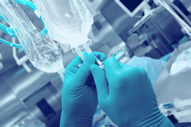 Pra Cego Ver: fotos de sudok1/iStock Banco de Imagens mostra a mão de um profissional da área da Saúde ajustando uma embalagem de soro/medicação