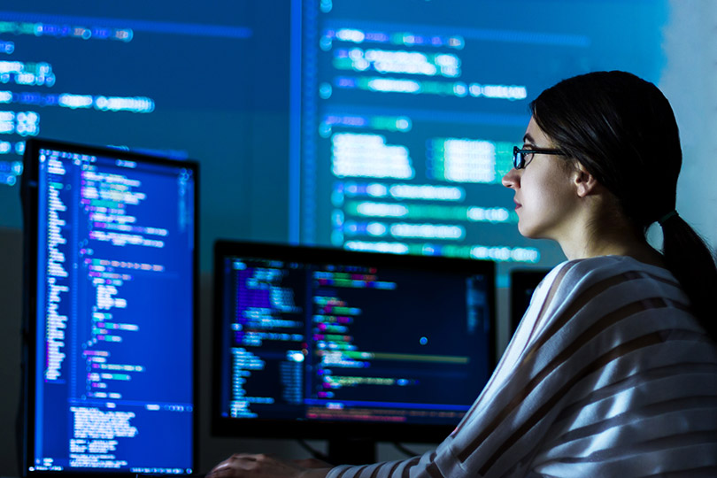 Pra Cego Ver: Foto de monstArrr_/iStock Banco de Imagens mostra programadora de TI em frente a monitores de computadores.