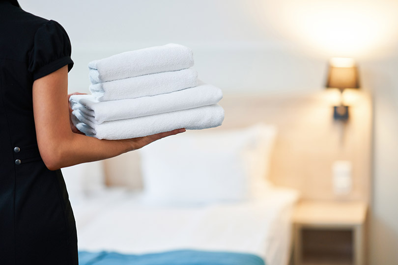 Pra Cego Ver: foto de macniak/iStock Banco de Imagens mostra uma camareira segurando toalhas e ao fundo uma cama de hotel.