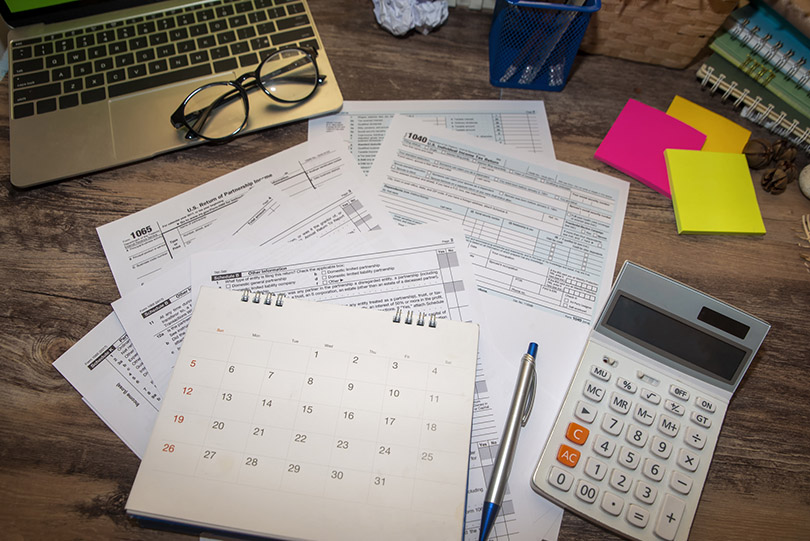 Pra Cego Ver: foto de Pra-chid/iStock Banco de Imagens mostra documentos relacionados ao Imposto de Renda, calculadora e notebook sobre mesa
