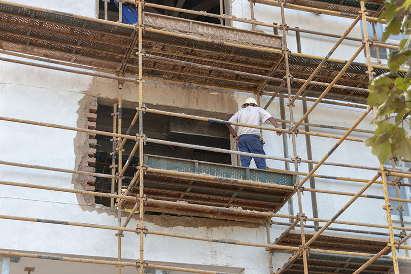 Pra Cego Ver: foto de kendalswart/depositphotos mostra um trabalhador, em pé, sobre um andaime, instalado na parede de um prédio.