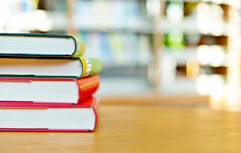 Fotografia de quatro livros empilhados em cima e uma mesa, em primeiro plano. Ao fundo, estantes de uma biblioteca, em desfoque.