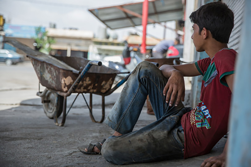 Pra Cego Ver: foto de Unicef mostra menino sentado no chão, com o rosto virado para o outro lado. Ele usa roupas sujas e aparenta cansaço. Olha para um carrinho de mão e para adultos que estão trabalhando.