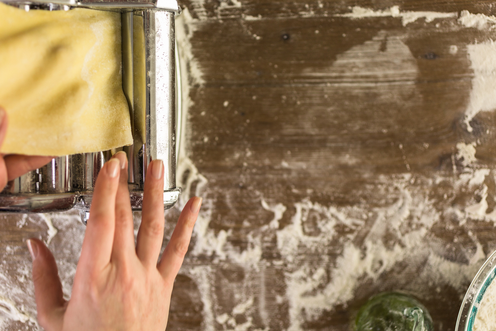 A imagem mostra uma mão feminina operando uma máquina de massas em uma mesa suja de farinha.