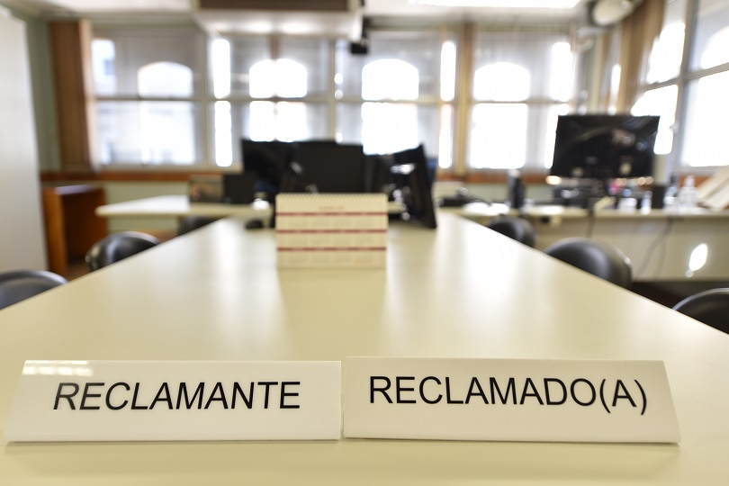 Foto de uma mesa de sala de audiências mostrando a placa "reclamante" de um lado e "reclamado(a)" do outro.