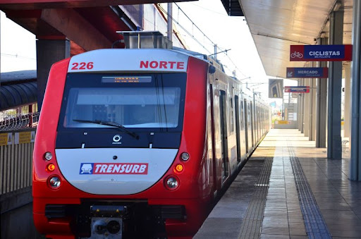 Foto que mostra um trem da trensurb parado em uma estação. A imagem mostra a frente do trem, à esquerda, e a plataforma de embarque e desembarque, à direita.