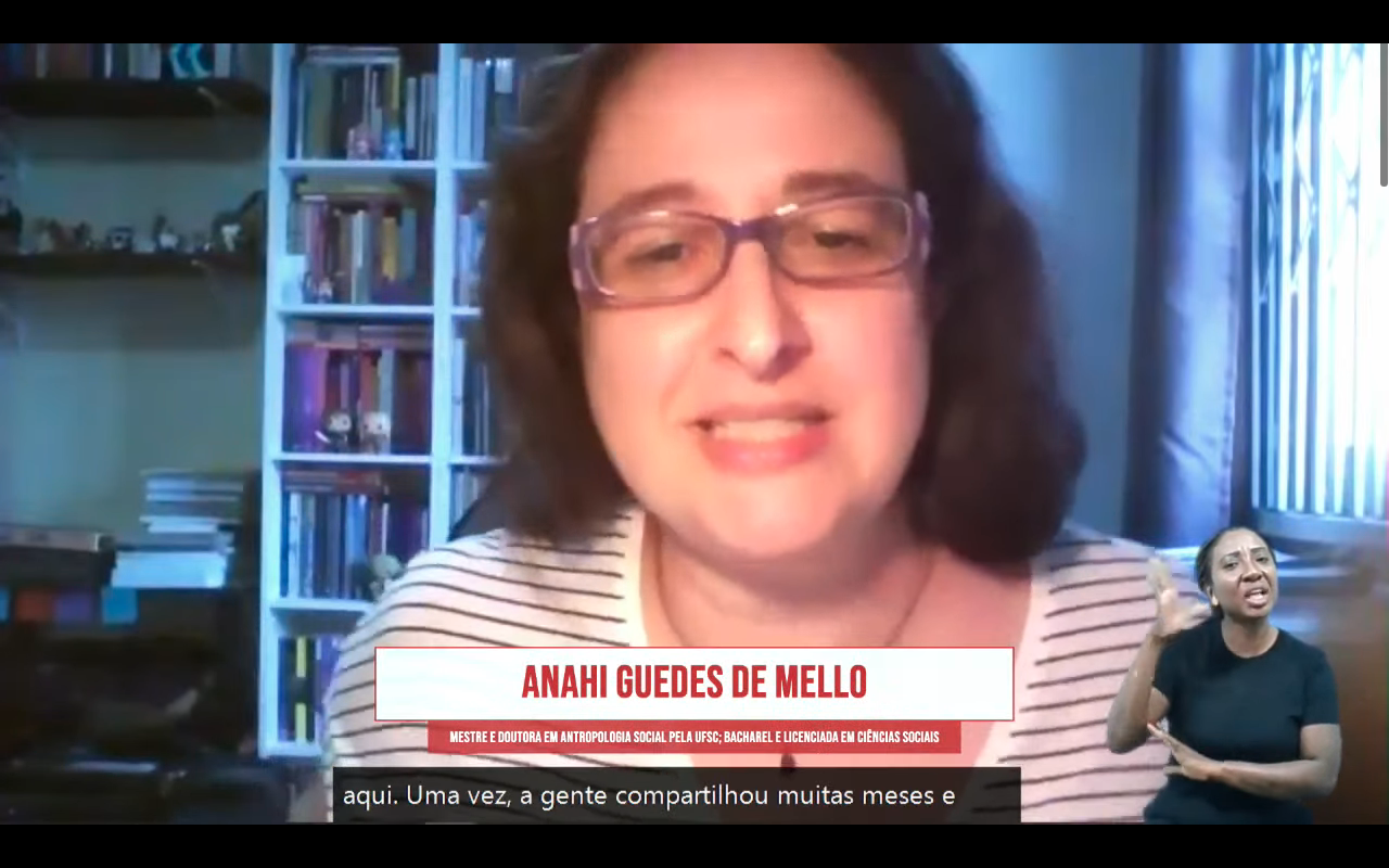 Foto de Anahi Guedes de Mello. É branca, usa óculos de armação roxa, camisa branca com listas pretas e um colar dourado. Ao fundo, uma estante com livros  e uma janela com a cortina fechada.