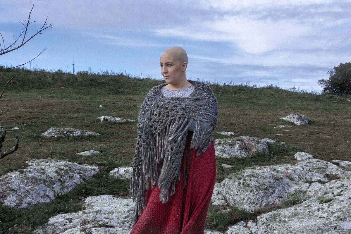 Mulher branca de cabelo raspado posa em um campo. Usa um xale cinza e uma saia vermelha, no estilo da personagem Ana Terra. Atrás ela aparece um gramado, rochas e galhos.