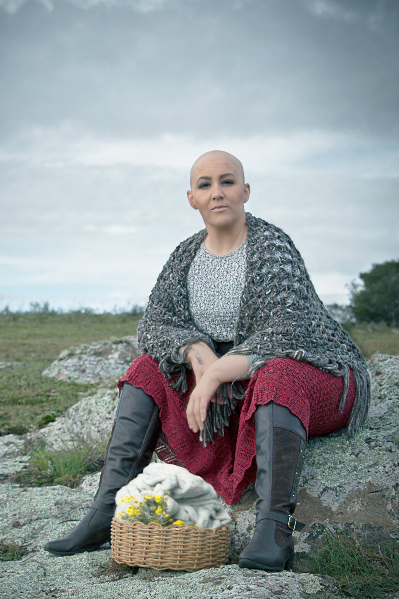 Mulher branca de cabelo raspado posa em um campo. Usa um xale cinza e uma saia vermelha, no estilo da personagem Ana Terra. Está sentada em uma pedra e abaixo dela há um cesto com flores.