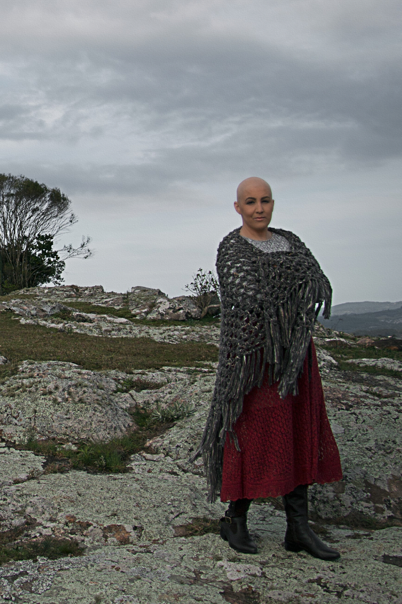 Mulher branca de cabelo raspado posa em um campo. Usa um xale cinza e uma saia vermelha, no estilo da personagem Ana Terra. Está de pé sobre um chão de pedras, com os braços cruzados. Ao fundo aparece um gramado e uma árvore.