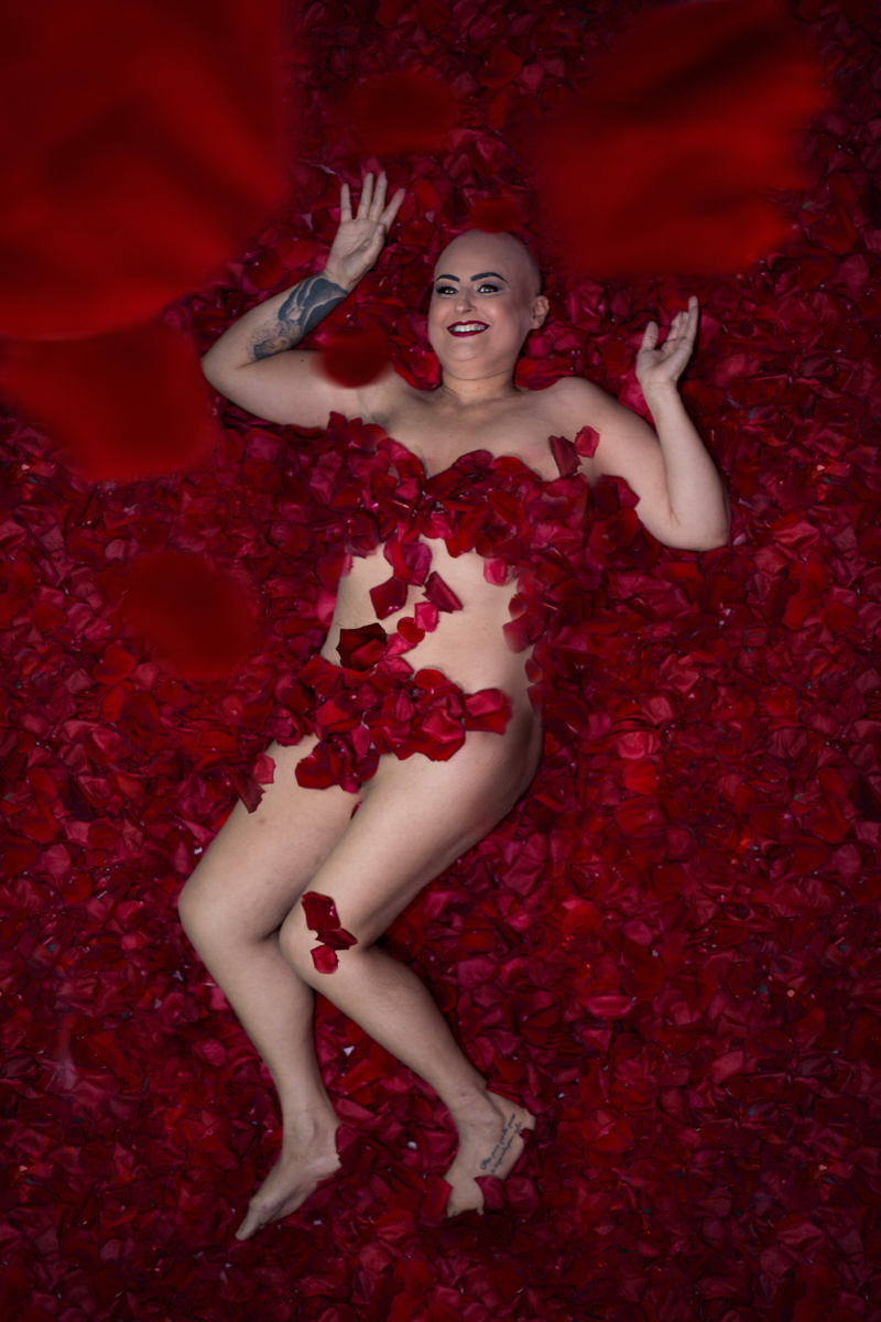 Mulher branca de cabelo raspado posa deitada sobre rosas vermelhas, semelhante à cena do filme Beleza Americana. Seu corpo está coberto apenas pelas rosas e ela está sorrindo para a câmera.