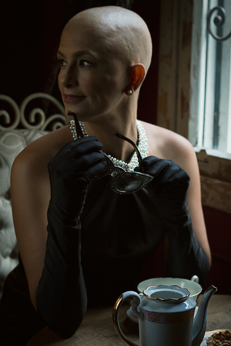 Mulher branca de cabelo raspado posa de perfil. Ela usa um colar de pérolas, um brinco de pérola, luvas escuras e segura um par de óculos escuros. na mesa em sua frente há um bule azul e uma xícara branca.