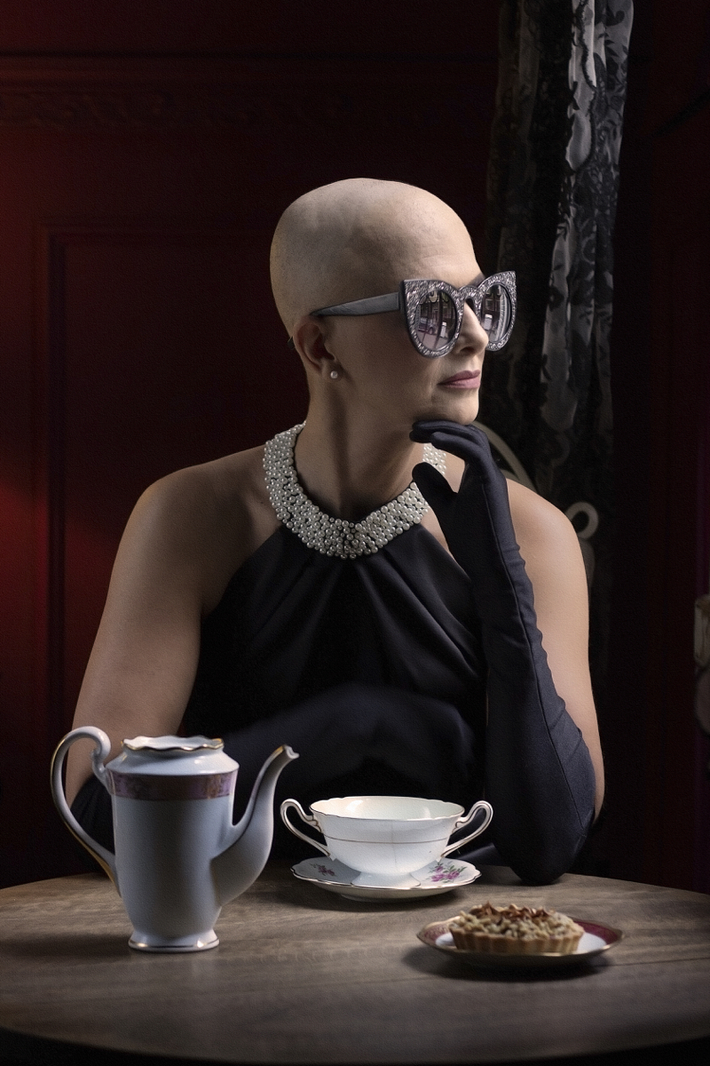 Mulher branca de cabelo raspado posa de perfil. Usaa um colar de pérolas, um brinco, luvas escuras e usa óculos escuros. Na mesa em sua frente há um bule azul e uma xícara branca.