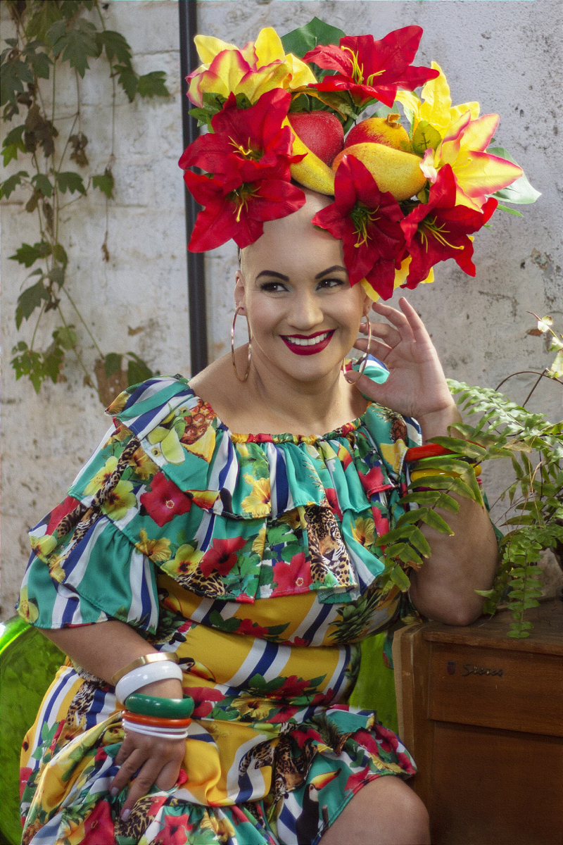 Mulher branca de cabelo raspado sorrindo de perfil. Usa um vestido colorido, com temas florais, e um arranjo artificial de flores e frutas na cabeça, remetendo à Carmen Miranda. Atrás dela há uma parede com folhagens.