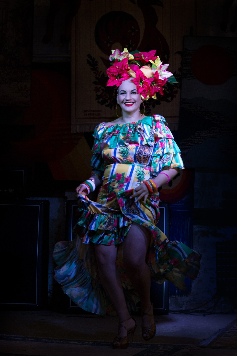 Mulher branca de cabelo raspado de pé, sorrindo, dançando em um palco. Usa um vestido colorido, com temas florais, e um arranjo artificial de flores e frutas na cabeça, remetendo à Carmen Miranda.